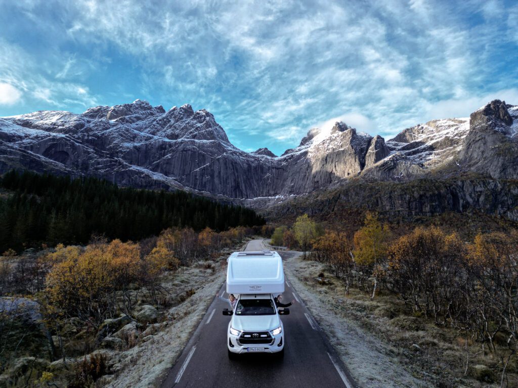 Truck Camper auf den Lofoten, mittig auf der Straße, im Hintergrund ein paar steile Berge. Aus dem Fenster des Truck Campers winken Sabine und Nico